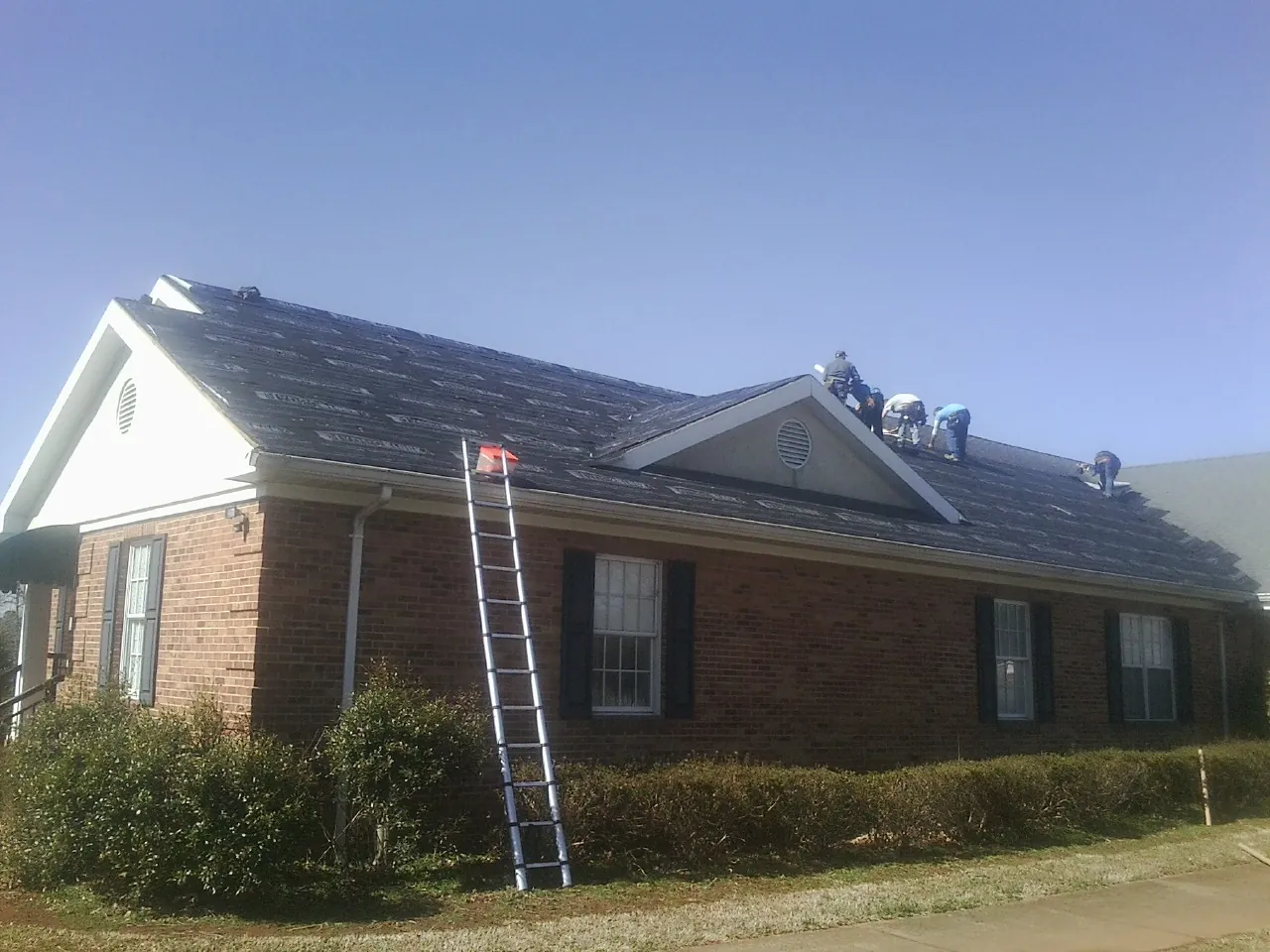 Crew tearing shingles off a brick ranch roof with a ladder set — Metal Roof Repair work by our crew in Spragueville