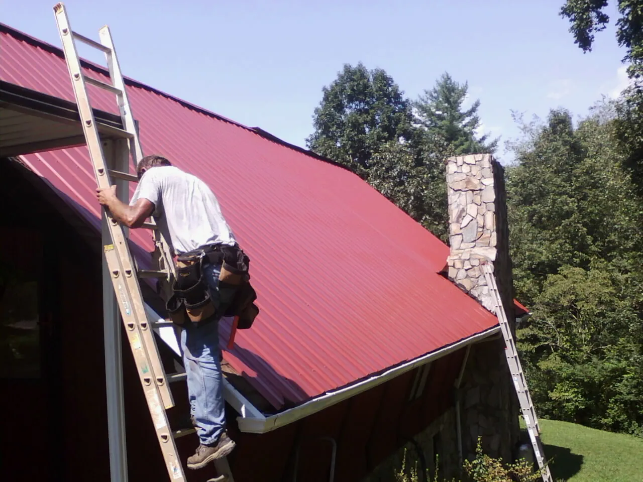Roofer on a ladder beside a red metal roof and stone chimney — Soffit & Fascia Repair by Roofing Brothers in Spragueville
